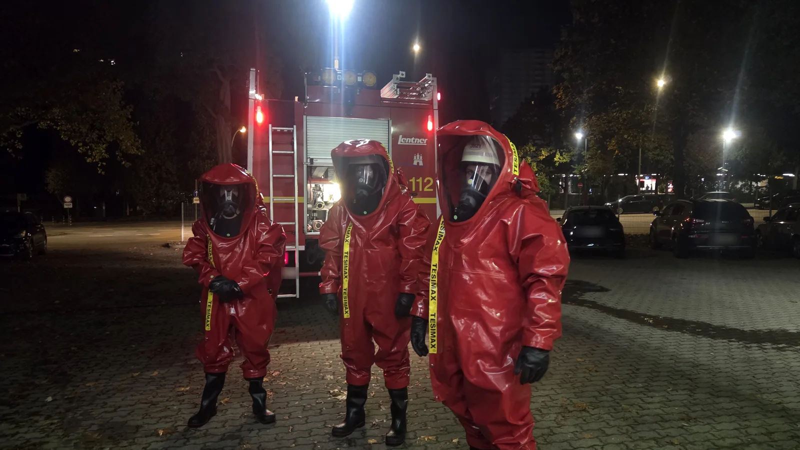 Drei Einsatzkräfte in roten Chemikalienschutzanzügen vor einem Löschfahrzeug
